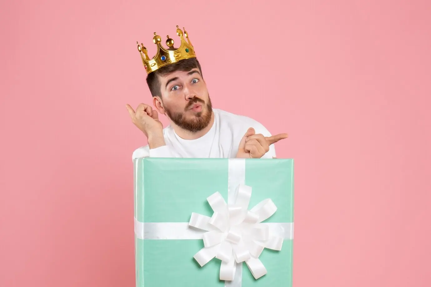 Front view of a young man inside a gift box with a crown against a pink wall